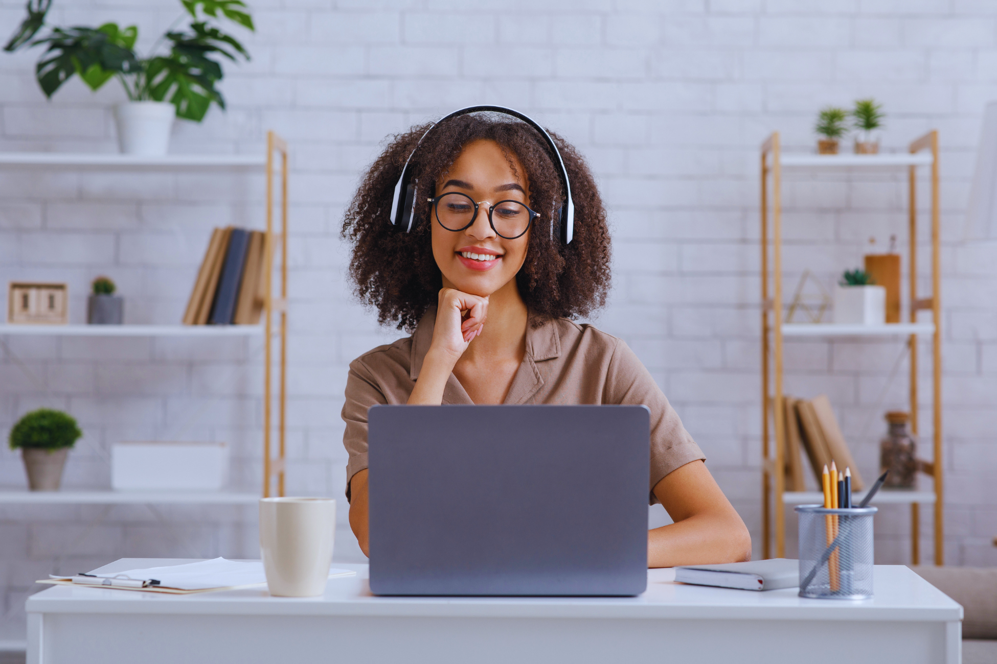 A woman wearing headphones sits at a desk, smiling at her laptop screen. Surrounded by office supplies, a coffee mug, and shelves with plants and books, she enjoys her bright, modern workspace focused on educação and learning.