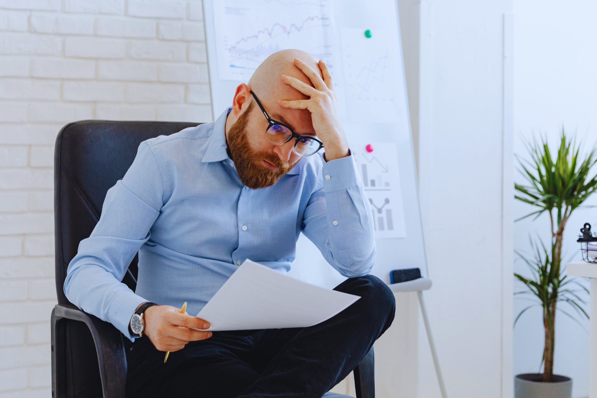 A man with glasses sits in a chair holding papers about the reforma da previdência and a pencil, looking stressed with his hand on his forehead. A whiteboard with charts and a potted plant are visible in the background.