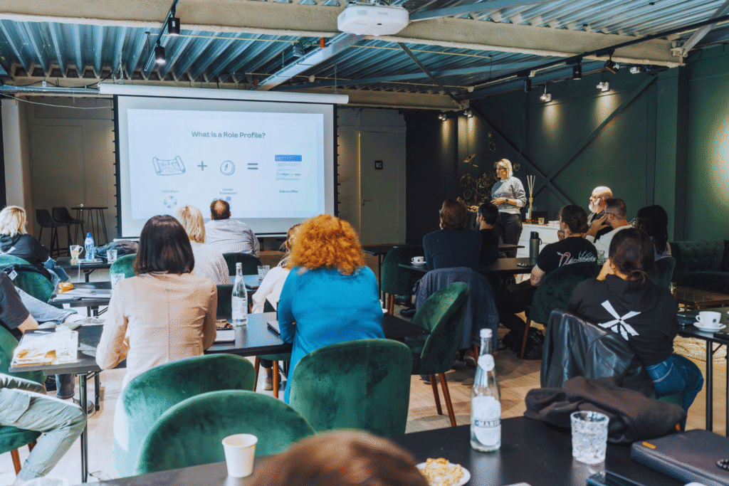 As pessoas assistem a uma apresentação em uma sala moderna e bem iluminada. Um palestrante fica ao lado de uma tela de projetor que exibe "What is a Role Profile?" (O que é um perfil de função?), compartilhando ideias que também podem ser encontradas no curso on-line. Os participantes se sentam em mesas com bebidas e papéis.