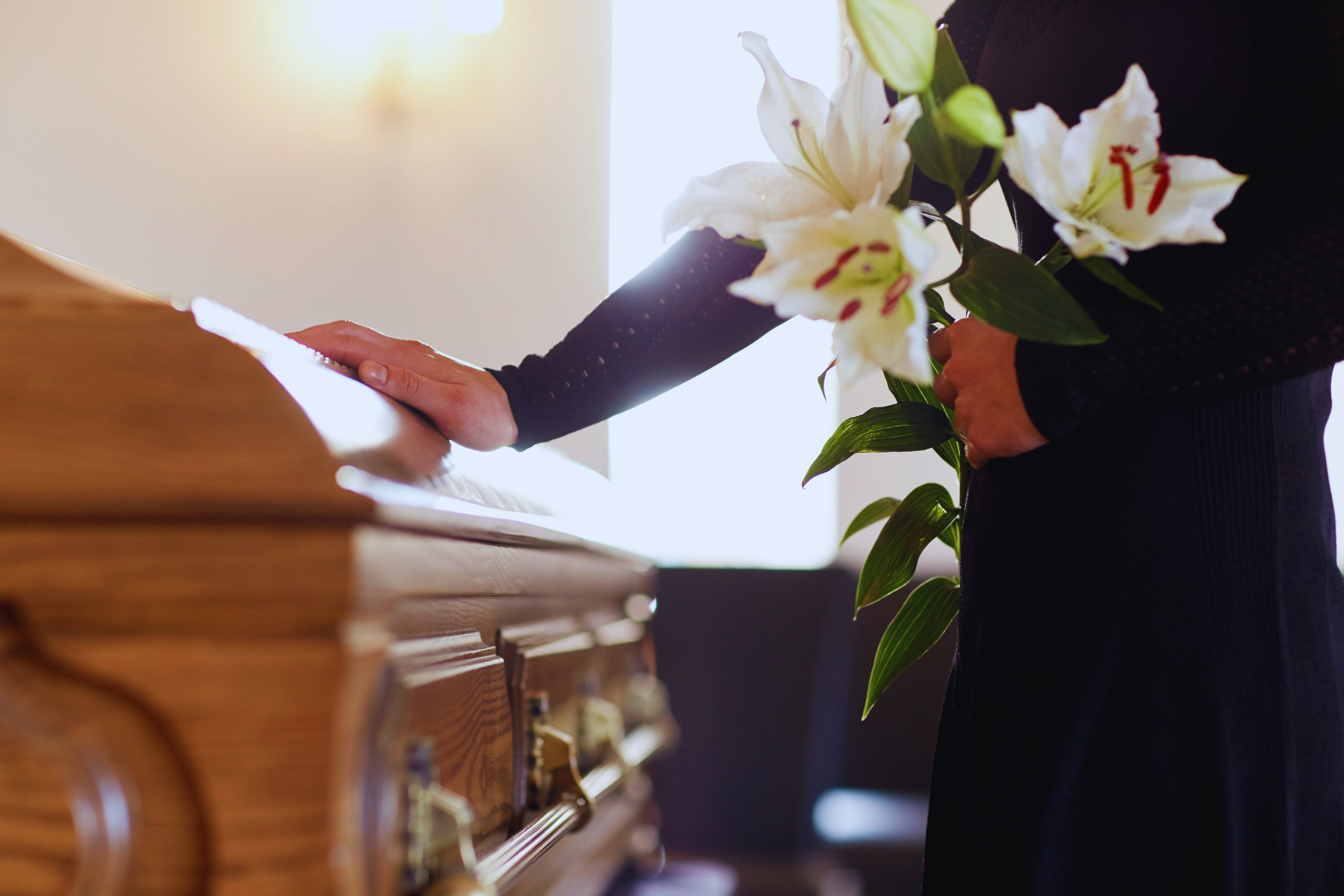 A person in black clothing stands beside a wooden casket, resting one hand on it and holding a bouquet of white lilies, symbolizing mourning at a funeral and the process of requesting Pensão por Morte.