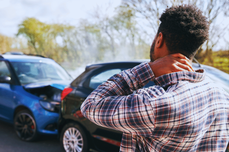 A man stands holding his neck in pain next to two cars that have collided on a sunny day, with danos materiais visible on the front of one car and the back of the other. Trees and sky are in the background.