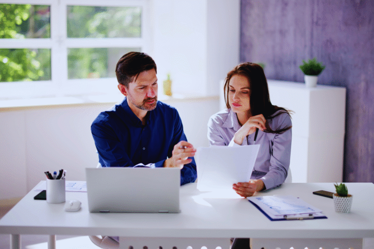A man and a woman sit at a desk with a laptop and documents, focused on reviewing a contrato de namoro. The man points to the paper the woman is holding as they engage in discussion. The office is bright with natural light and minimal decor.
