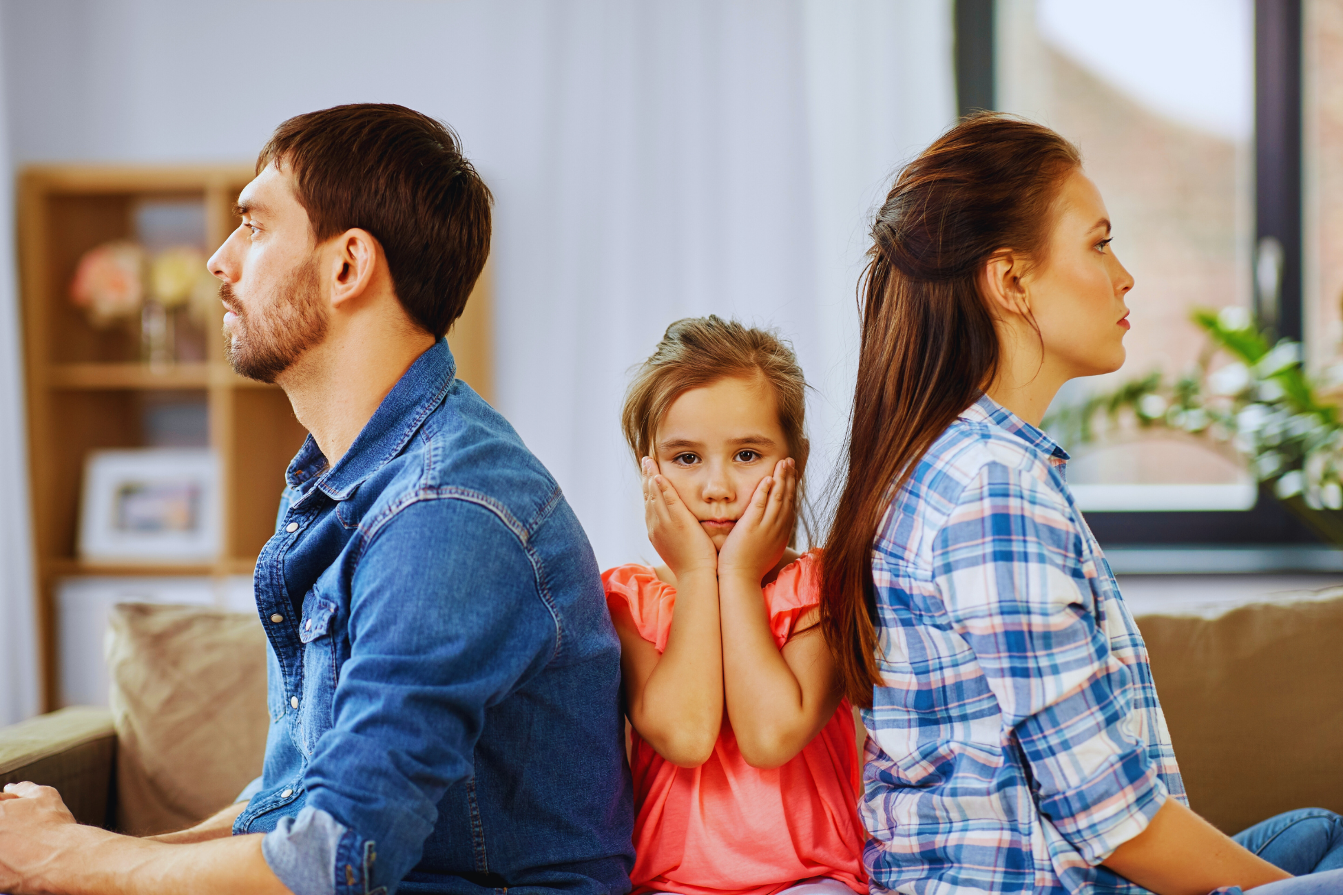 A young girl sits between two adults on a couch, holding her face with her hands and looking concerned, while the adults, facing away from each other, display signs of distress during a dissolução da união estável.