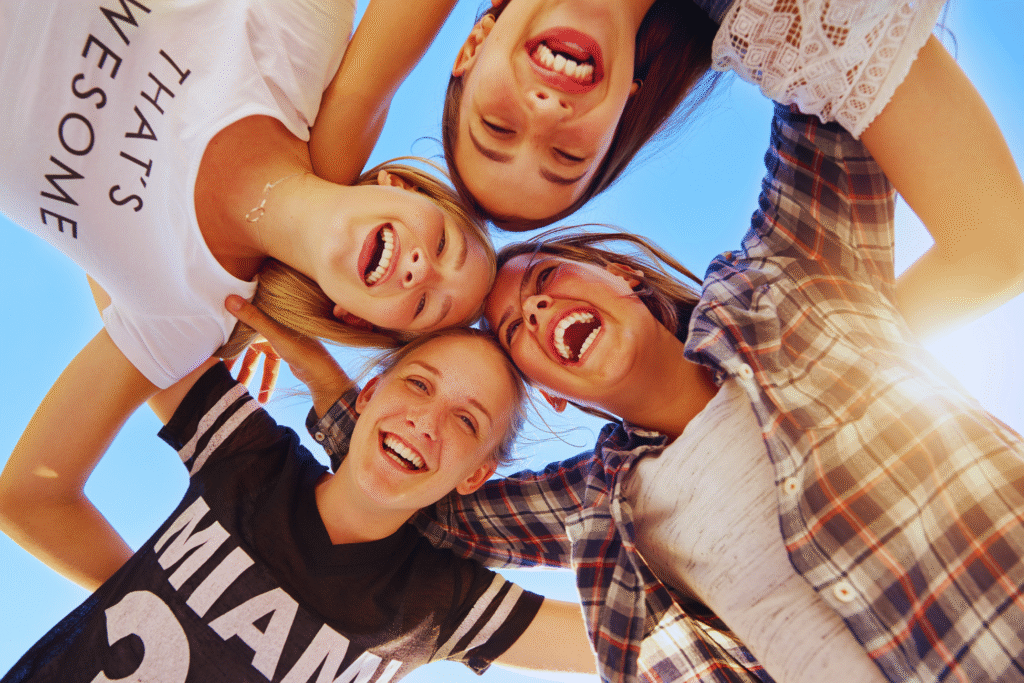 Four smiling teenagers stand in a circle, arms around each other, looking down at the camera with joyful expressions against a bright blue sky, celebrating their sense of amizade and emancipação.