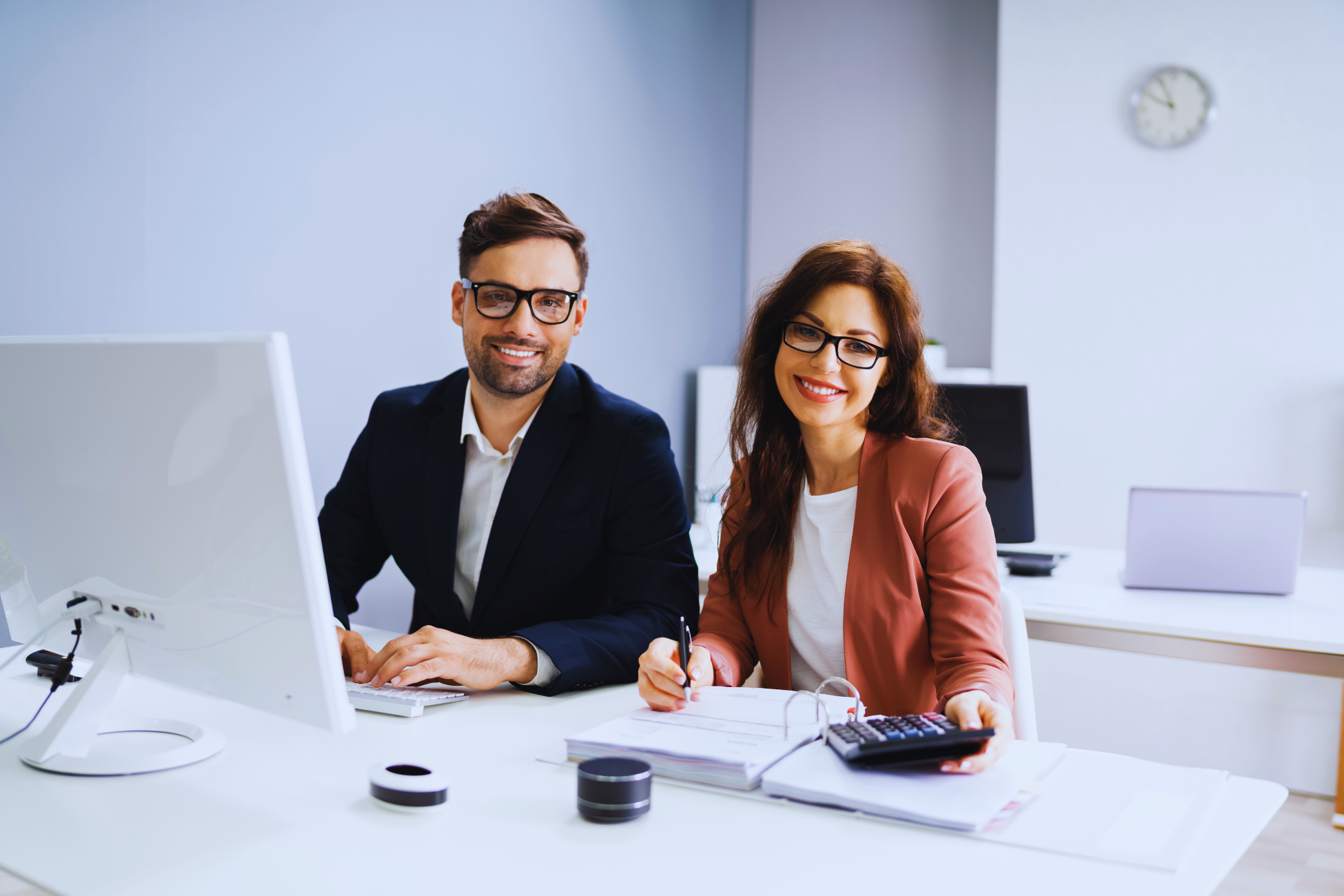 Two business professionals, a man and a woman, sit at a desk in a modern office, smiling at the camera. Surrounded by computers and notebooks, they appear ready to discuss financial reports or iniciar uma ação de exigir contas.