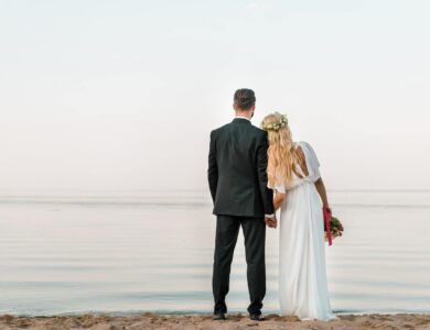 back view of wedding couple standing on beach with wedding bouquet and looking at sea