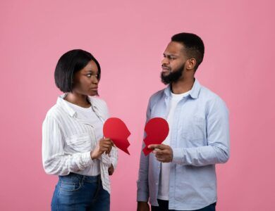 Displeased black guy and lady holding halves of torn paper heart on pink studio background. Divorce