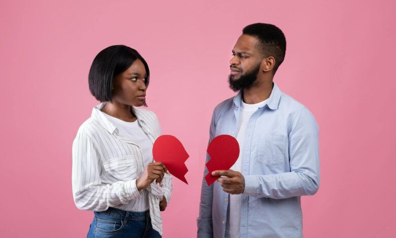 Displeased black guy and lady holding halves of torn paper heart on pink studio background. Divorce