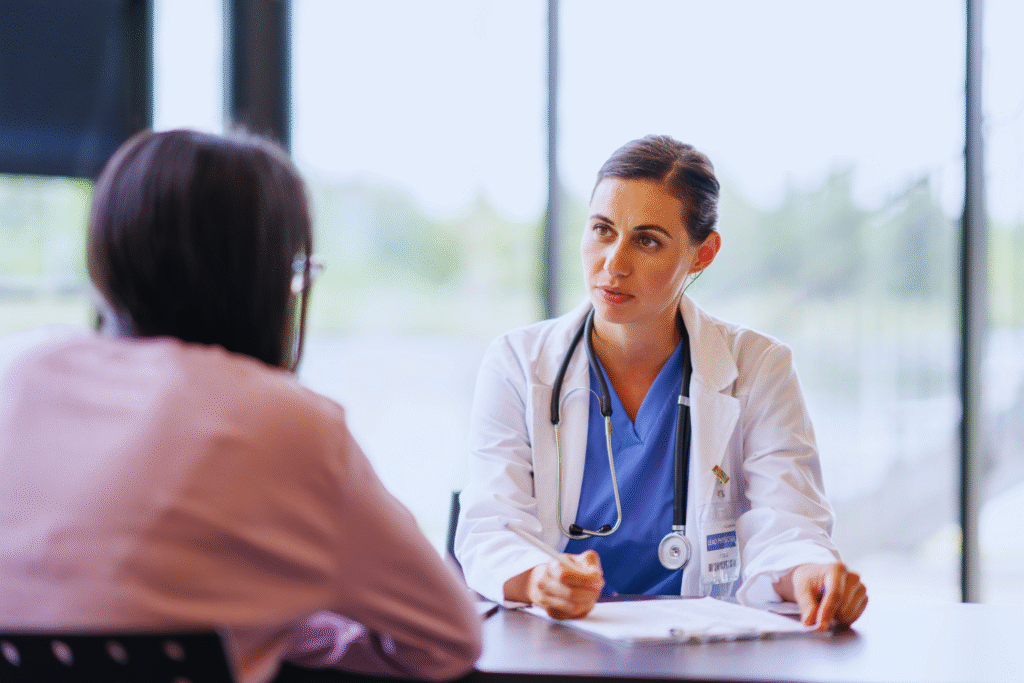 Uma médica de jaleco branco e estetoscópio discute sobre indenização contra o INSS com um paciente em uma mesa em uma sala bem iluminada com grandes janelas. A médica parece atenta e envolvida na conversa.