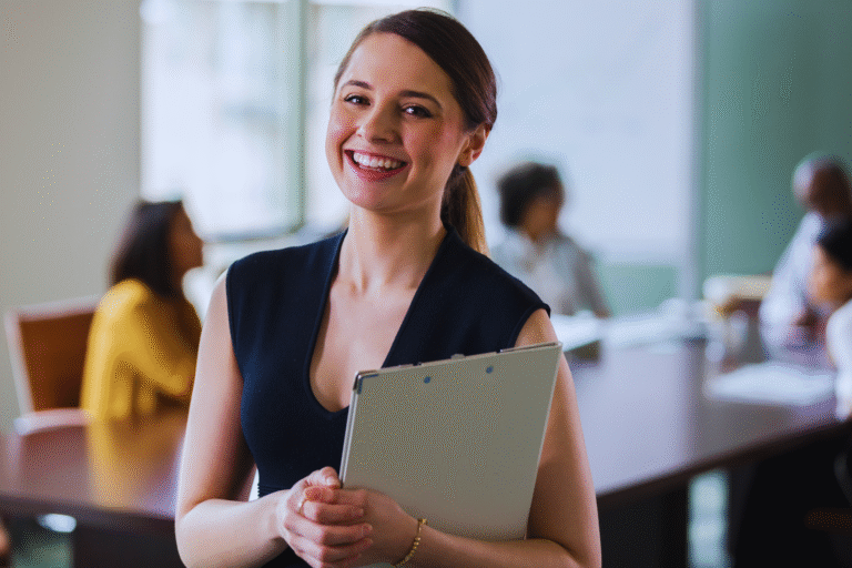 A smiling woman holding a folder stands in a modern office, possibly preparing a requerimento administrativo ou judicial para Isenção, while three people sit and talk at a conference table in the blurred background.