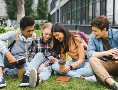 smiling and happy teenagers sitting on grass and looking at smartphone
