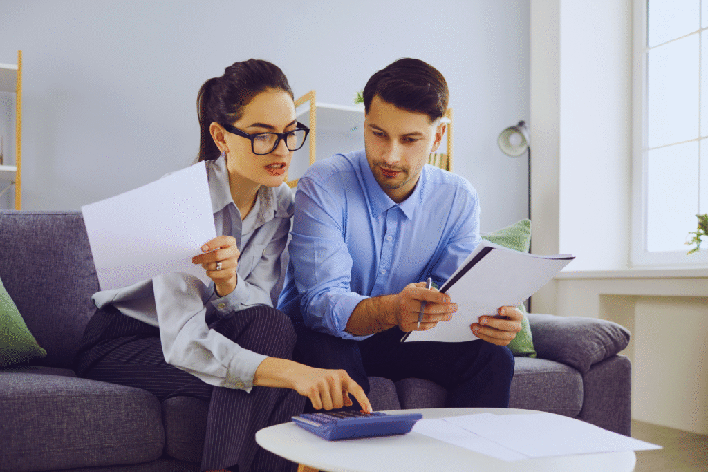 A woman and a man sit on a couch reviewing documents. The woman, holding papers, uses a calculator, while the man with a clipboard watches intently as they discuss como declarar empréstimo no Imposto de Renda.