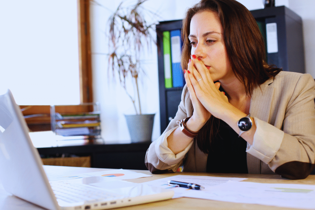 A woman in a beige blazer sits at a desk, looking thoughtfully at a laptop screen, possibly researching como evitar cair na malha fina. Papers, a pen, and a plant are visible in the background.
