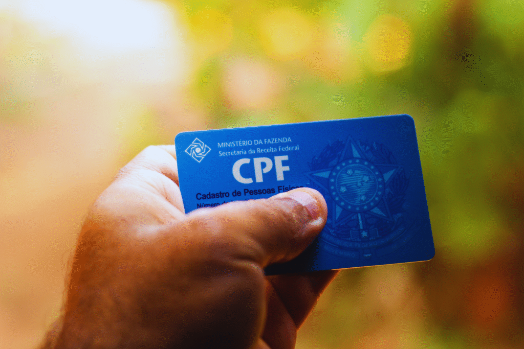 Uma mão segurando um cartão azul do CPF (Cadastro de Pessoas Físicas) brasileiro, essencial para declarar imposto de renda, com vegetação desfocada ao fundo.