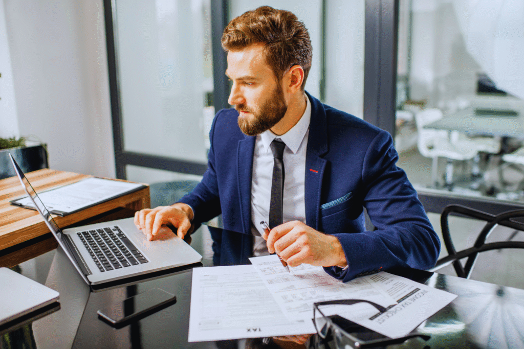 A man in a navy suit sits at a desk in an office, declarar imposto de renda on his laptop with tax documents and pen in hand. A smartphone and paperwork are on the desk, and large windows are in the background.