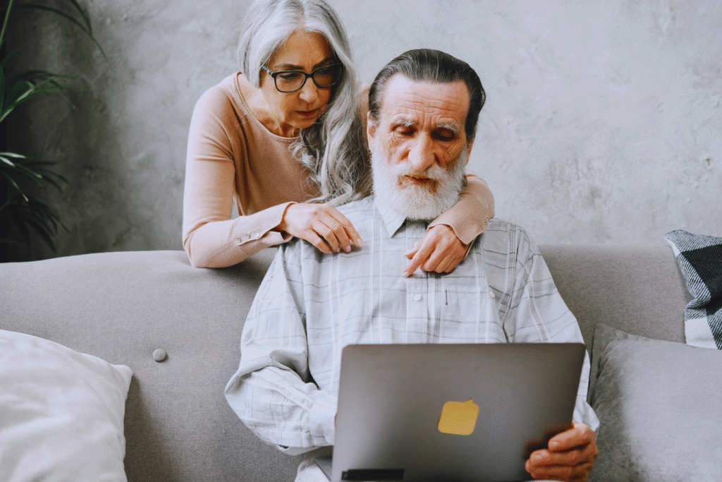 An older man with a gray beard sits on a couch using a laptop to check revisão decorrente de erro de cálculo do INSS, while a woman with long gray hair stands behind him, looking at the screen over his shoulder with her hands resting gently on his shoulders.