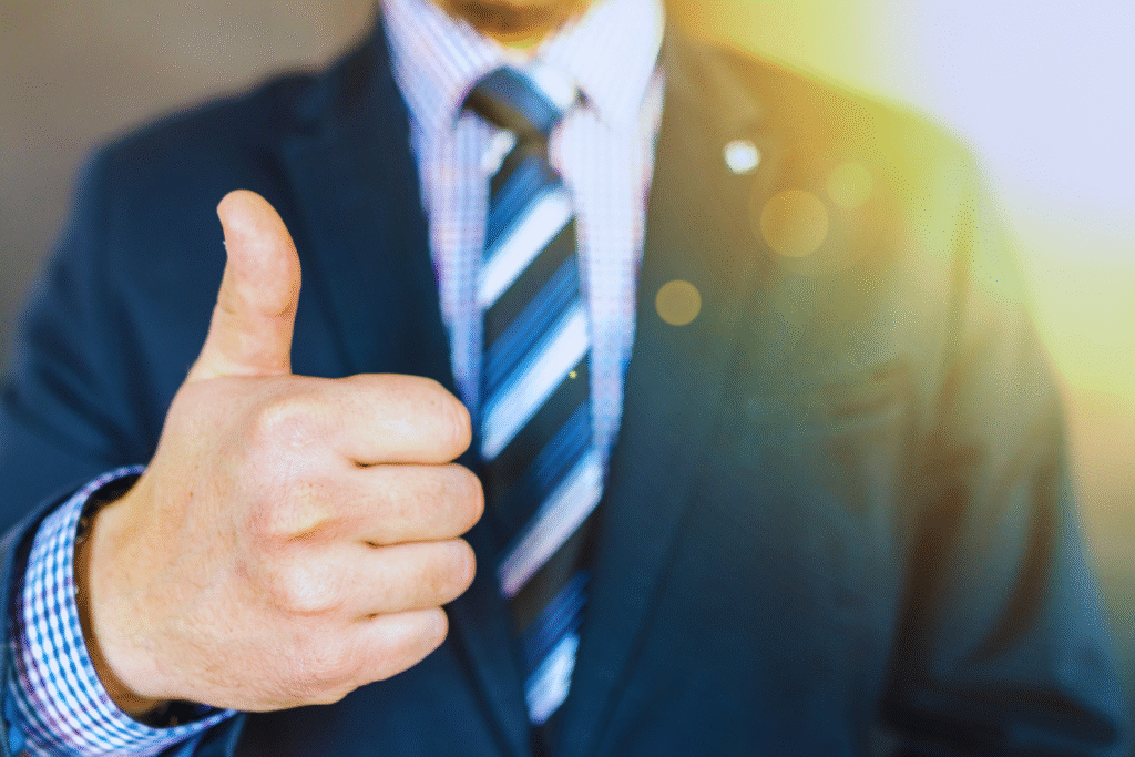 Um homem de terno e gravata faz um gesto de polegar para cima, com a luz do sol brilhando ao fundo - simbolizando a recuperação e a positividade após um Acidente do Trabalho. O foco está em sua mão, enquanto seu rosto está fora de quadro.