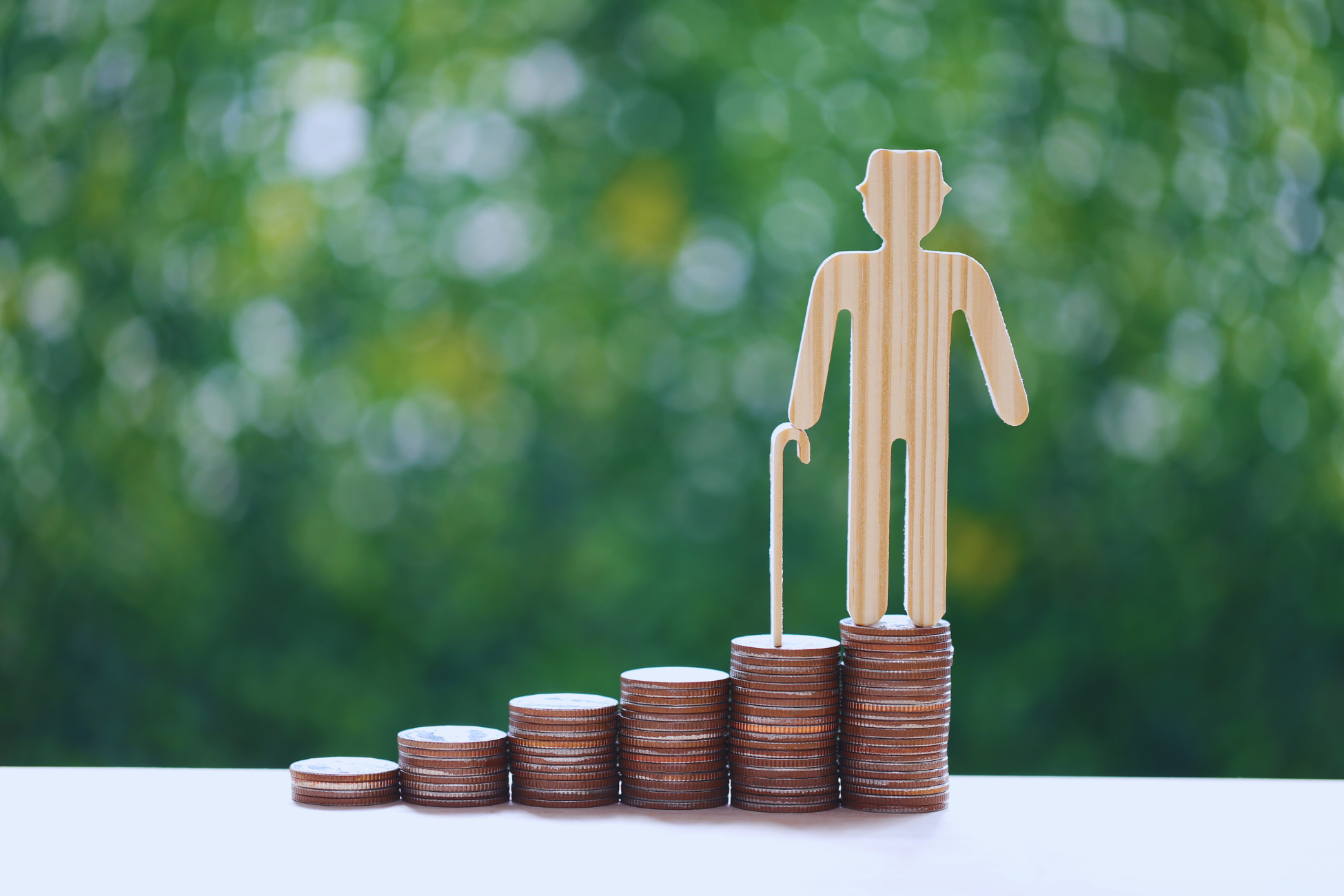 A wooden figure of an elderly person with a cane stands on stacked coins arranged in ascending order, symbolizing retirement savings, pensão vitalícia por acidente, or financial growth for old age, with a blurred green background.