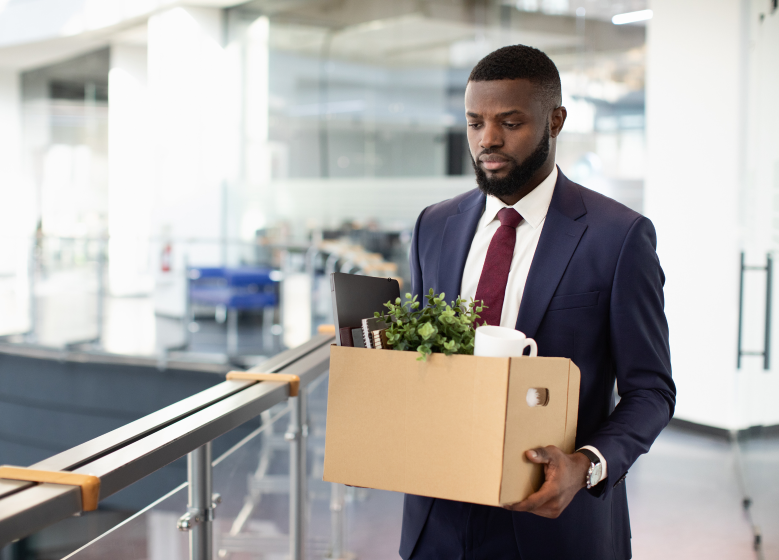 Um homem de terno carrega uma caixa de papelão com material de escritório e uma planta, caminhando por um moderno prédio de escritórios com uma expressão séria - uma imagem que captura o momento do abandono de emprego.