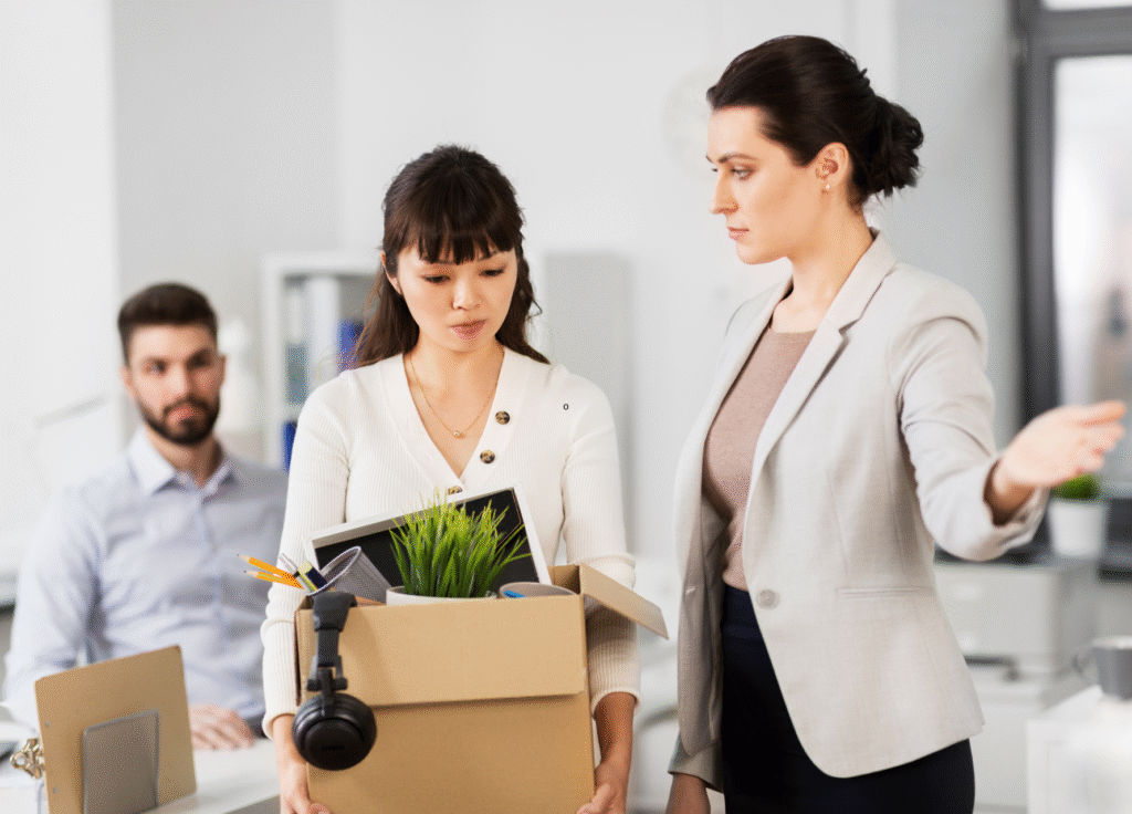 Uma mulher segurando uma caixa com itens pessoais olha para baixo com tristeza após uma demissão por justa causa, enquanto outra mulher em trajes profissionais gesticula em direção à saída; um homem está sentado ao fundo observando a cena em um ambiente de escritório.