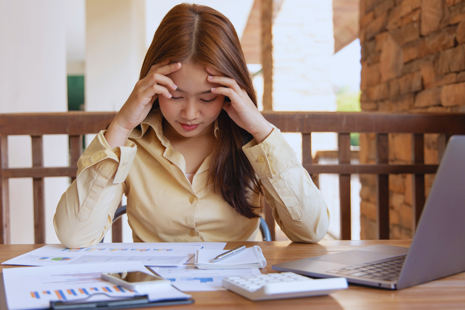A woman sits at a table, looking stressed with her hands on her temples. Surrounded by documents, a laptop, a calculator, and a phone, she appears overwhelmed, likely from long hours and dealing with horas extras at work.