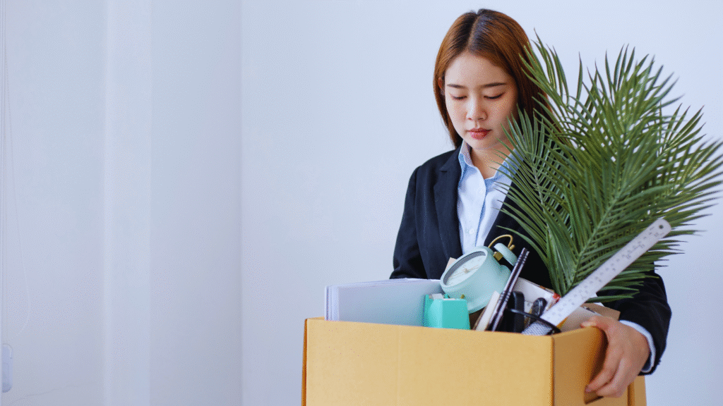Uma mulher em um terno preto olha para baixo com tristeza enquanto segura uma caixa de papelão com material de escritório, um vaso de planta e itens pessoais, destacando o impacto do abandono de emprego quando ela deixa seu emprego.