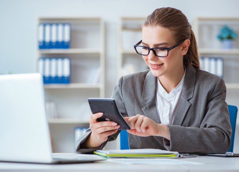 Uma mulher de terno cinza e óculos está sentada em uma mesa, sorrindo enquanto usa uma calculadora, possivelmente mostrando como calcular hora extra. Um laptop, documentos e pastas são visíveis no fundo claro e organizado do escritório.