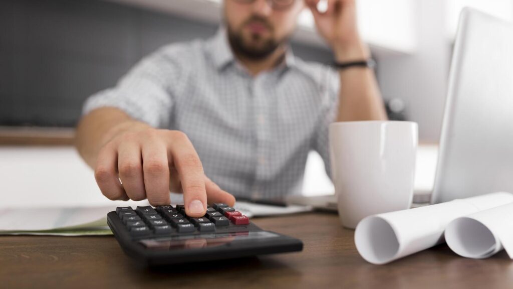 Uma pessoa vestindo uma camisa xadrez usa uma calculadora em uma mesa com uma caneca de café, dois papéis enrolados e um laptop ao fundo, parecendo pesquisar como calcular insalubridade para trabalho ou cálculos financeiros.
