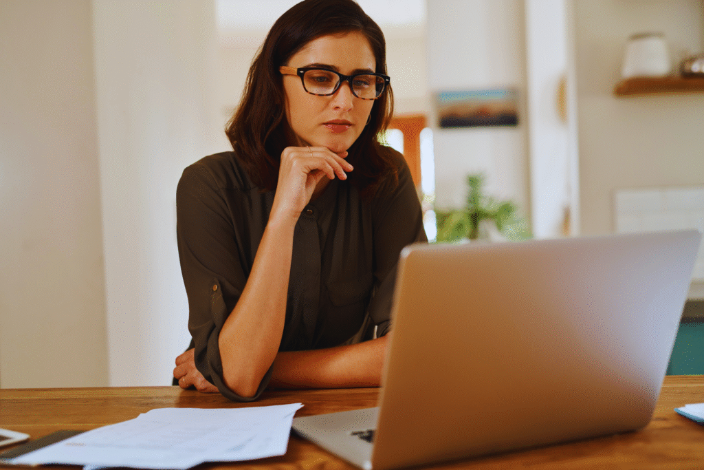 Uma mulher de óculos está sentada em uma mesa, olhando atentamente para seu laptop enquanto pesquisa como calcular o seguro desemprego. Papéis e um smartphone estão sobre a mesa de madeira, sugerindo um ambiente de trabalho doméstico ou de escritório.