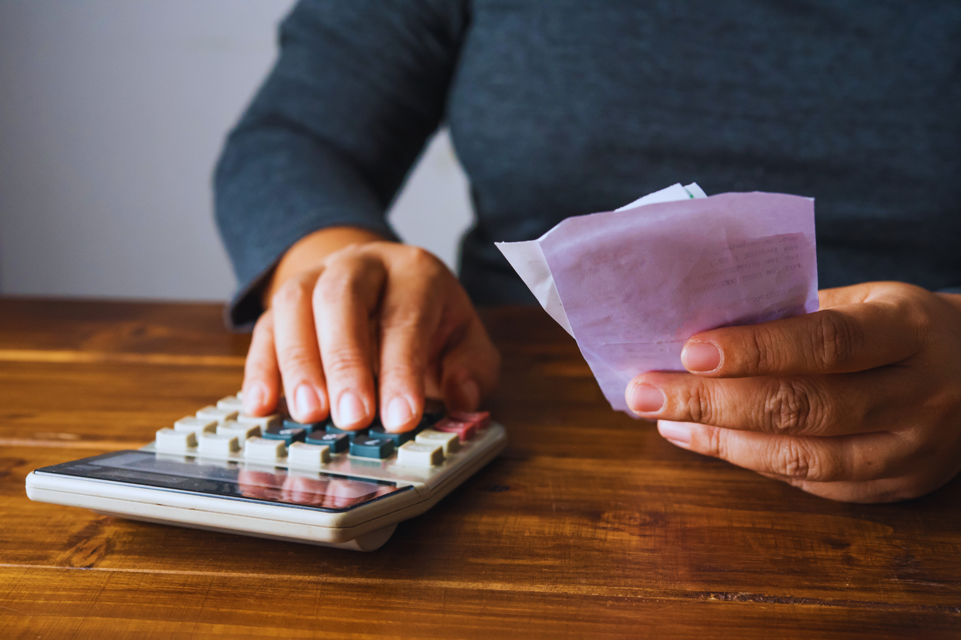 A person sitting at a wooden table uses a calculator with one hand while holding several receipts in the other, appearing to manage finances or learn como calcular o seguro desemprego.