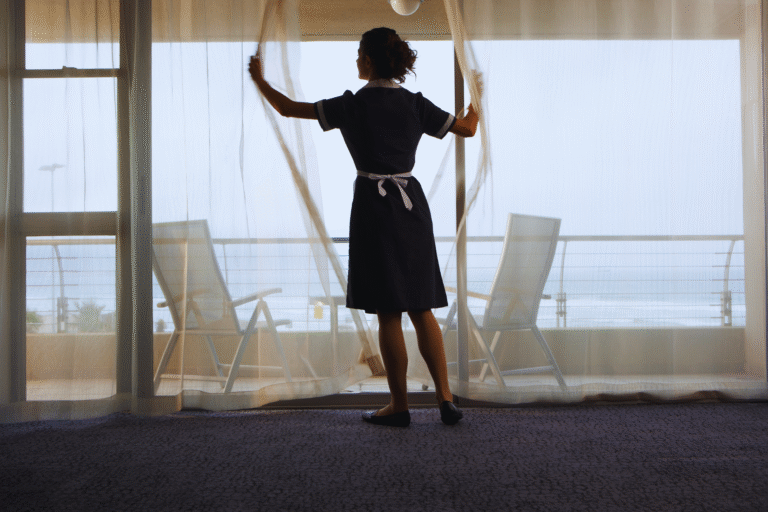 A person in a cleaning uniform stands by a large window, holding open sheer curtains. It raises the question: empregada doméstica tem direito a seguro desemprego? Two empty chairs and a railing are visible on the balcony outside, with an overcast sky beyond.