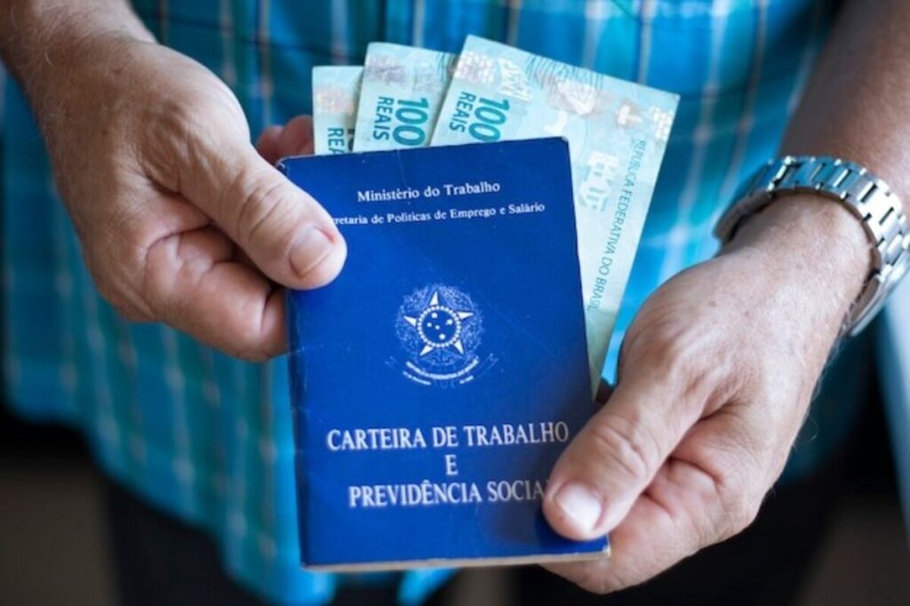 Uma pessoa segura uma Carteira de Trabalho e Previdência Social brasileira, duas cédulas de 100 reais e veste uma camisa xadrez azul, simbolizando o acesso aos direitos e benefícios dos trabalhadores, como o FGTS.