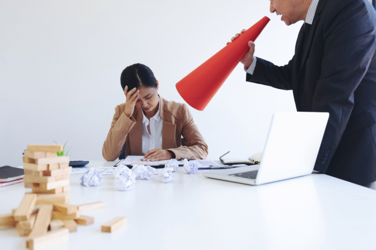 Um homem grita através de um megafone vermelho para uma mulher estressada sentada em uma mesa desordenada, destacando o assédio moral no ambiente de trabalho em uma cena tensa de escritório com papéis espalhados e uma torre de Jenga derrubada.