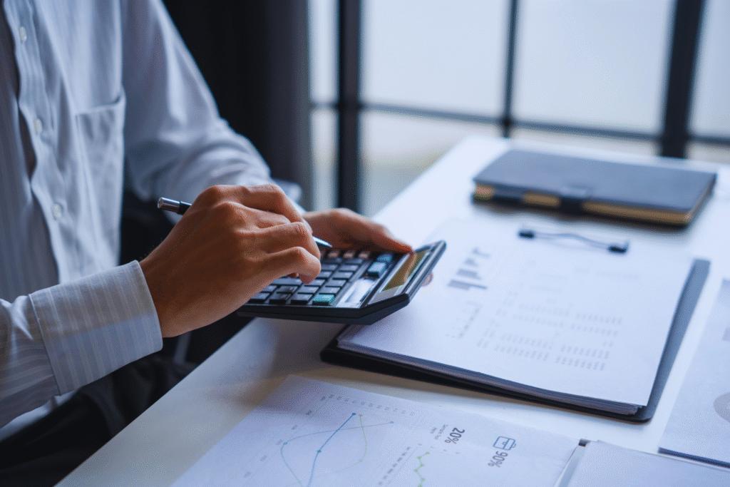 Uma pessoa de camisa social usa uma calculadora em uma mesa com documentos financeiros, gráficos e um caderno, analisando detalhes sobre aviso prévio indenizado por uma janela com luz natural.