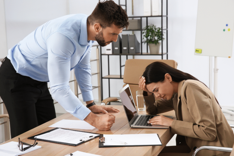 Um homem de camisa azul se posiciona sobre uma mulher sentada em uma mesa, levantando a voz - um exemplo de tipos de assédio moral no trabalho. A mulher parece estressada, segurando a cabeça com a mão enquanto olha para o laptop.