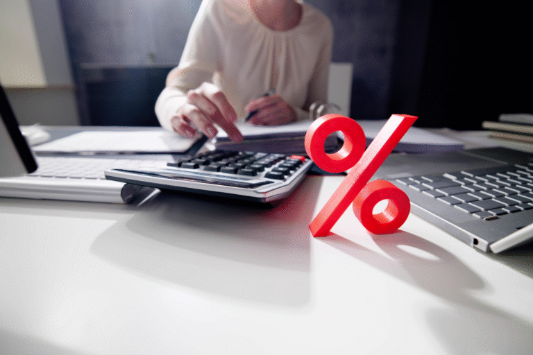 Uma pessoa usa uma calculadora em uma mesa com papéis, um teclado e um grande símbolo vermelho de porcentagem em primeiro plano, ilustrando como calcular o décimo terceiro ou realizar cálculos de taxas de juros.