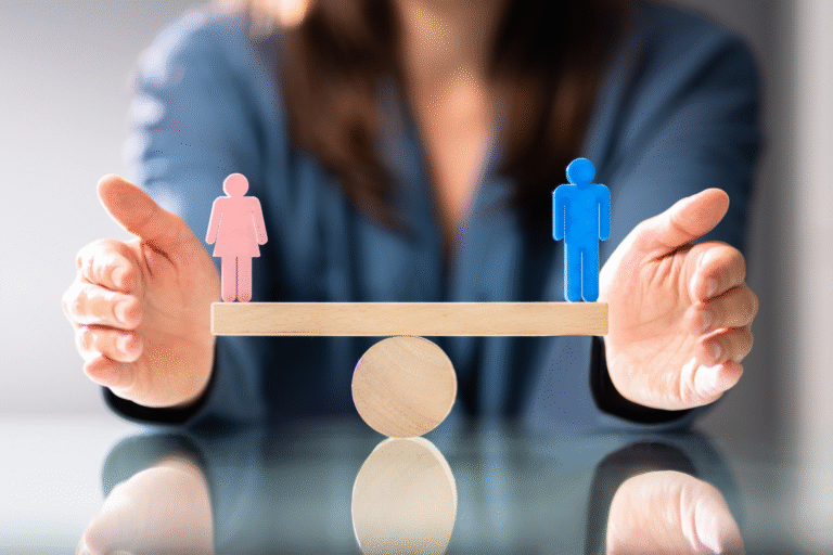 Uma pessoa segura uma gangorra de madeira equilibrada com figuras femininas cor-de-rosa e masculinas azuis em cada extremidade, simbolizando a igualdade de gênero e a importância da lei de igualdade salarial.