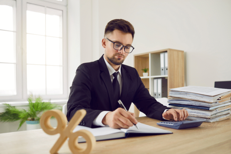 Um homem de terno está sentado em uma mesa escrevendo em um caderno, com uma pilha de documentos e uma calculadora ao seu lado - talvez revisando se o décimo terceiro salário tem desconto de INSS. Um sinal de porcentagem de madeira está sobre a mesa em primeiro plano.