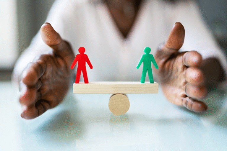 A person’s hands frame a seesaw balancing two wooden human figures, one red and one green, symbolizing o que é equiparação salarial—equality or balance between two people.
