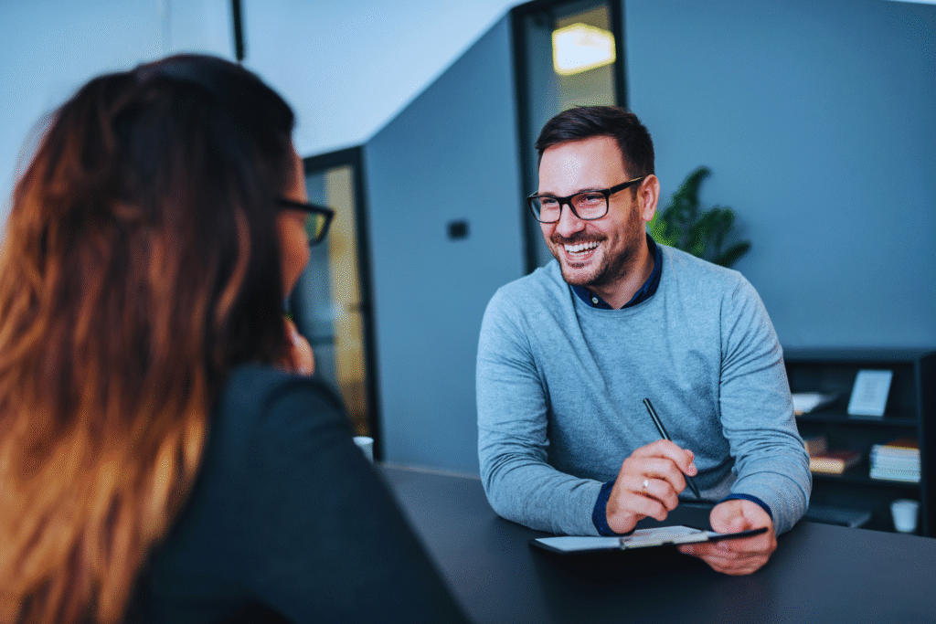 Duas pessoas estão sentadas em uma mesa em um ambiente de escritório. O homem, usando óculos e um suéter cinza, sorri enquanto segura uma caneta e um caderno, discutindo estabilidade no emprego com a mulher à sua frente.