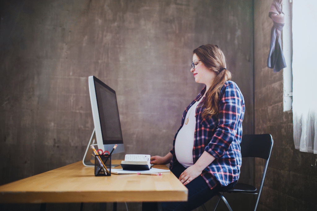 Uma mulher grávida, segura pela estabilidade gestante CLT, está sentada em uma mesa trabalhando em um computador em uma sala com pouca iluminação e uma parede de concreto. Ela usa óculos, camisa xadrez e calça jeans, e tem cabelos compridos puxados para trás.