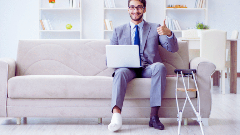 A man in a suit sits on a couch with a laptop on his lap, smiling and giving a thumbs up. With one foot bandaged and elevated—perhaps em estabilidade auxílio doença—his crutches rest nearby. A modern bookshelf and table complete the scene.