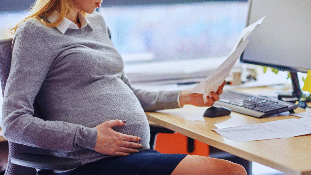 Uma mulher grávida senta-se em uma mesa de escritório, segurando documentos e apoiando a mão na barriga - talvez considerando questões como "quanto tempo dura a estabilidade da gestante" enquanto revisa a papelada em seu suéter cinza e saia azul-marinho.