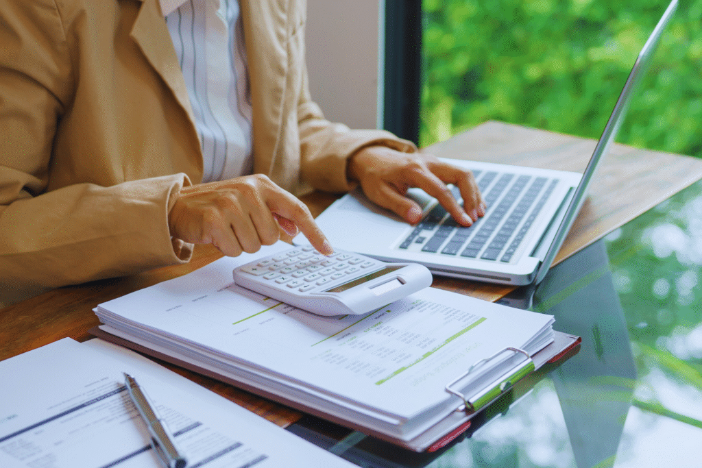 A person in a tan blazer uses a calculator and types on a laptop at a desk covered with financial documents, charts, and notes related to abono pecuniário de férias, with a window showing greenery in the background.
