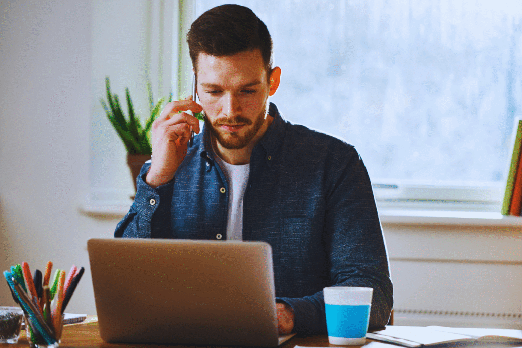 Um homem com barba senta-se em uma mesa usando seu laptop para uma consulta de processo trabalhista enquanto fala ao telefone. Uma xícara, canetas coloridas e uma planta estão sobre a mesa, com uma janela e livros ao fundo.