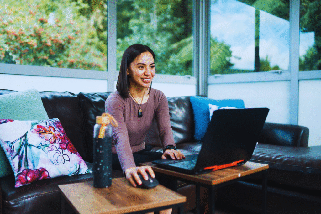 Uma mulher está sentada em um sofá usando um laptop e um mouse sobre pequenas mesas, sorrindo enquanto lê sobre a lei sobre trabalho híbrido. Ela veste um top lilás, com uma garrafa de água por perto e grandes janelas atrás dela mostrando a vegetação do lado de fora.
