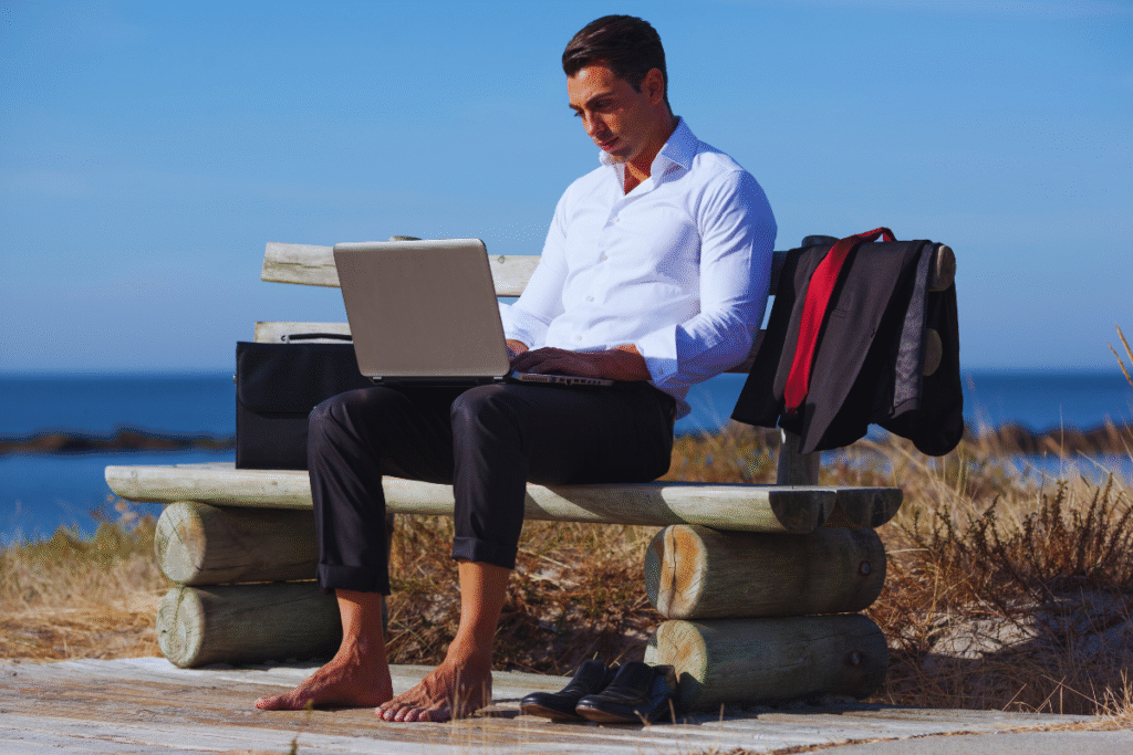Um homem em trajes de negócios senta-se descalço em um banco de madeira à beira-mar, trabalhando em um laptop - uma imagem que captura o trabalho híbrido e remoto. Seus sapatos e sua jaqueta estão sobre o banco, com uma pasta próxima e água azul clara ao fundo.