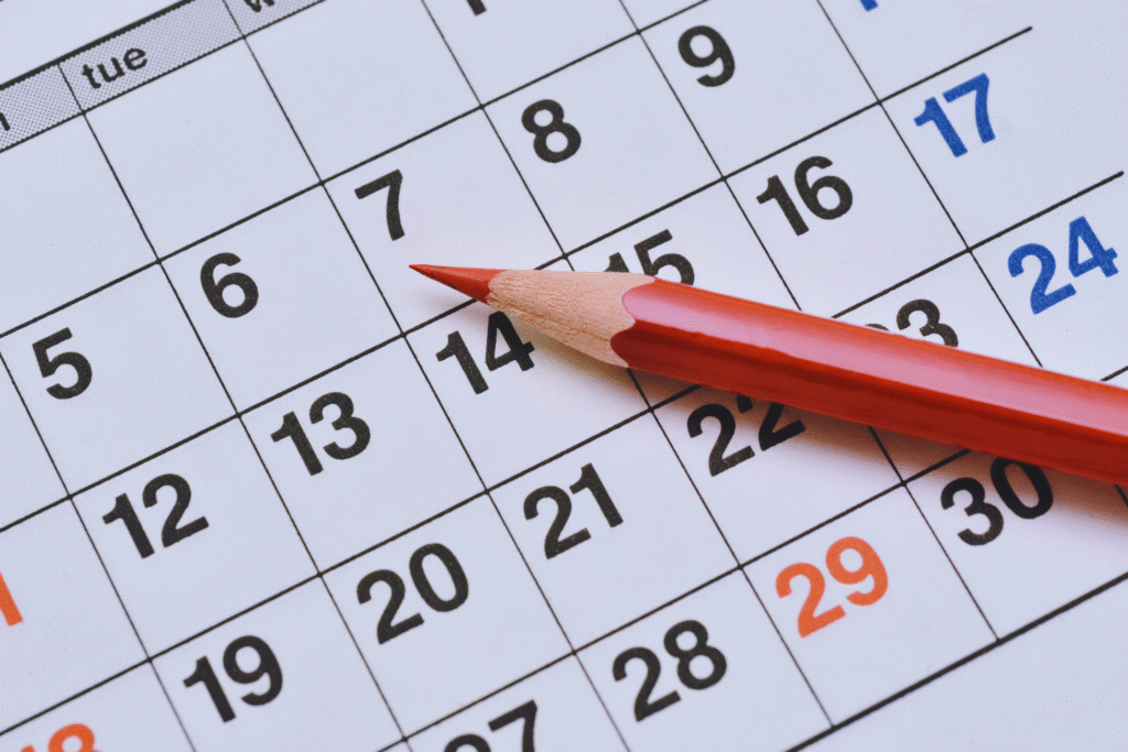A red pencil rests on a calendar, pointing at the 14th day of the month—a useful tool for understanding como funciona a escala 6x1. The dates are printed in black, with Sundays in blue and the 29th highlighted in orange.