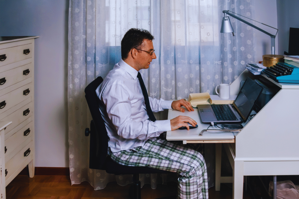 Um homem abraça o teletrabalho em um escritório doméstico, usando camisa social, gravata e calça de pijama xadrez enquanto trabalha em seu laptop em meio a papéis, um caderno, uma caneca e uma luminária.