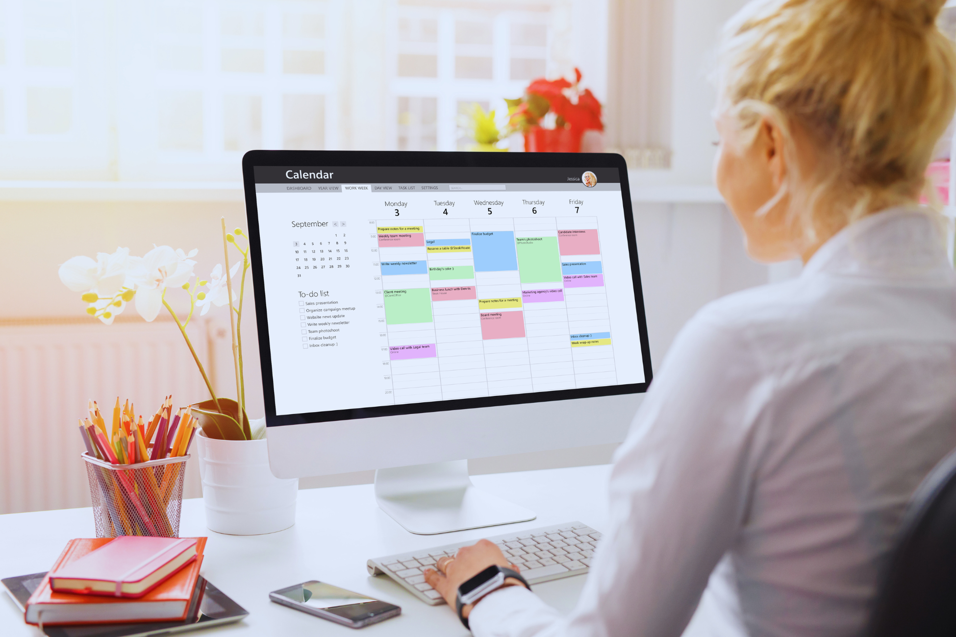 A woman sits at a desk using a computer with a digital calendar displayed on the monitor, showing a busy weekly schedule and tips on como fazer escala de trabalho. The workspace has flowers, notebooks, and sunlight coming through a window.